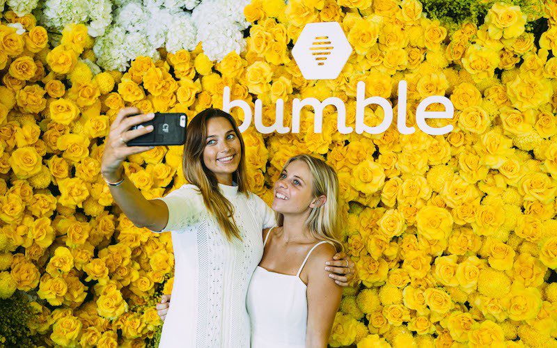 Two women taking a selfie at a wall of yellow flowers that says 'bumble'